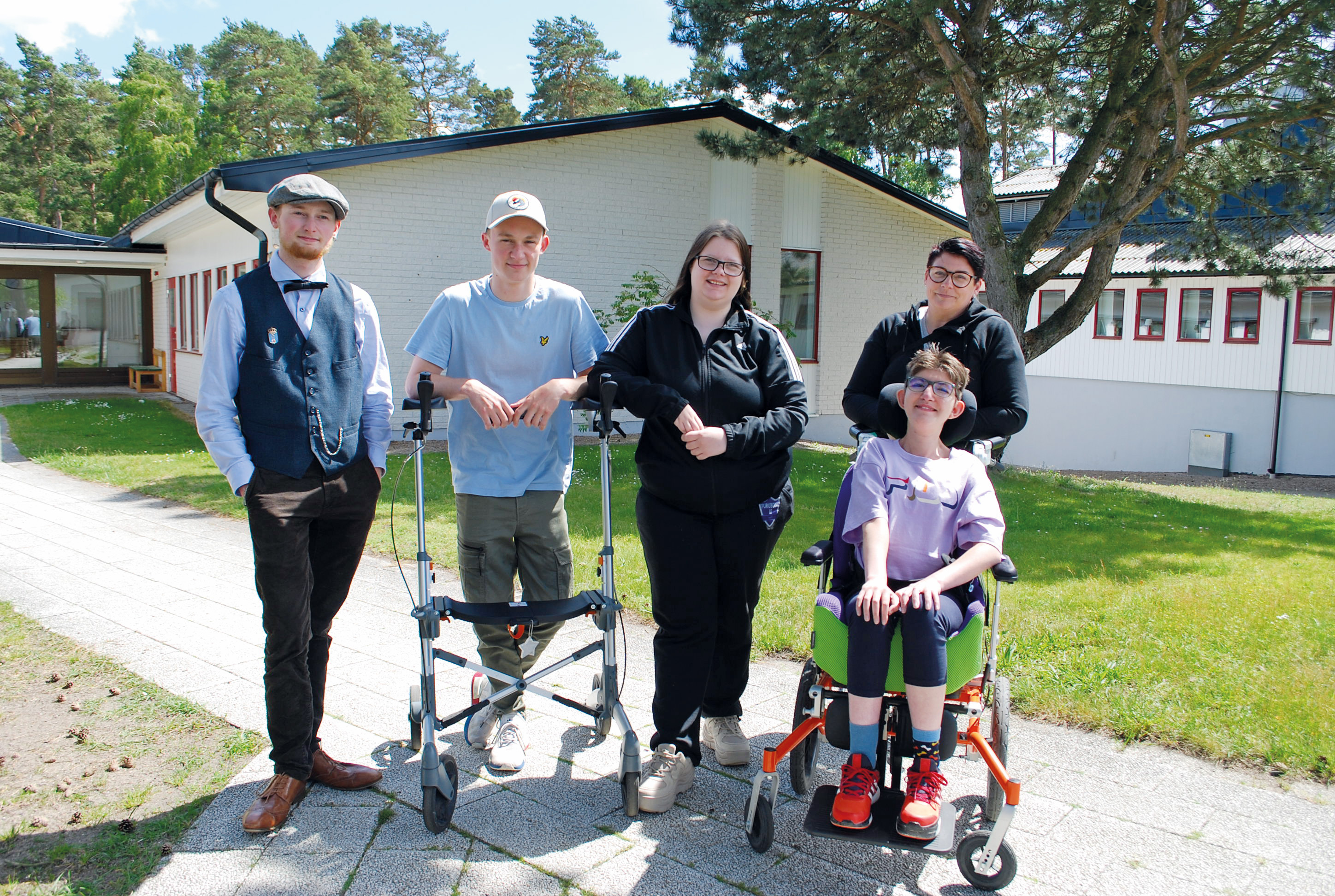 Fyra personer som står och en som sitter i en rullstol utomhus. De är glada,